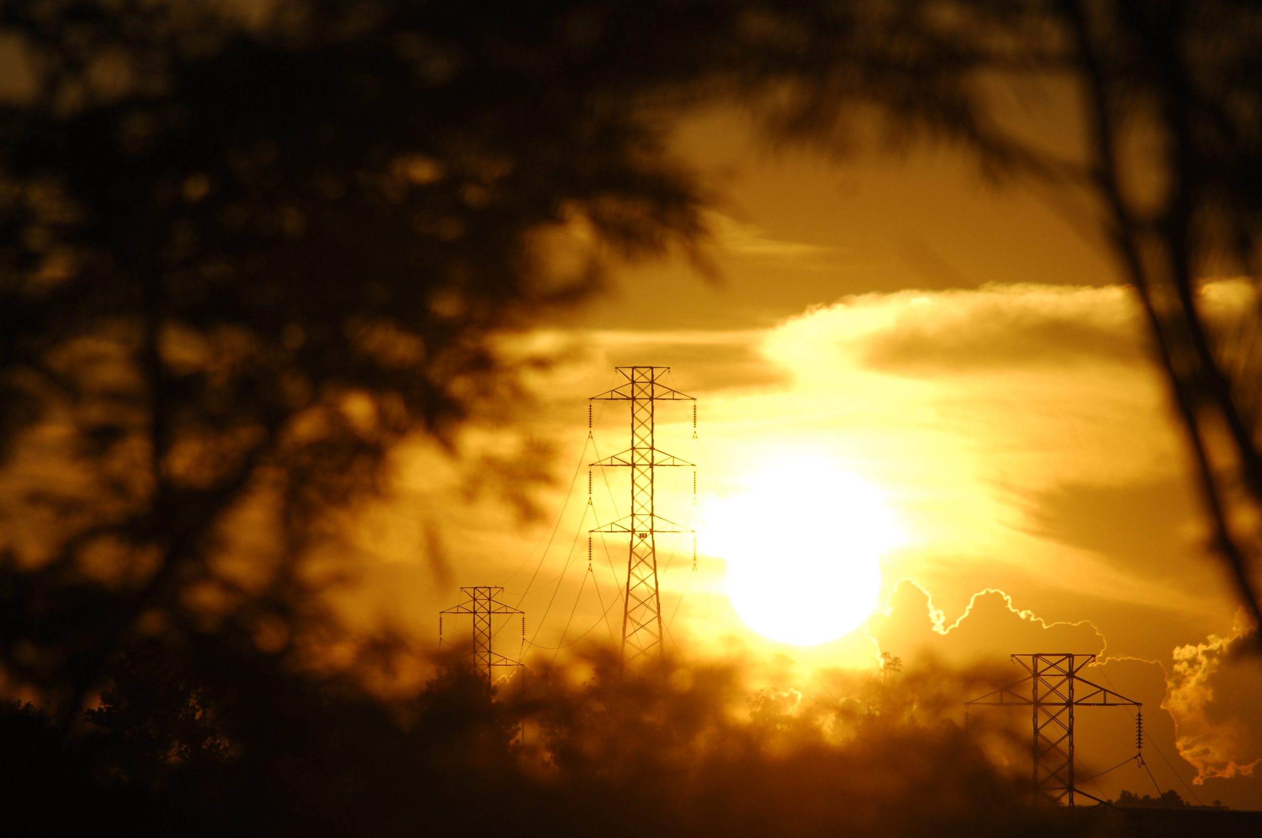 Hay una torre de transmisión de isa enmarcada en un paisaje que contiene un sol anaranjado de fondo y árboles desenfocados